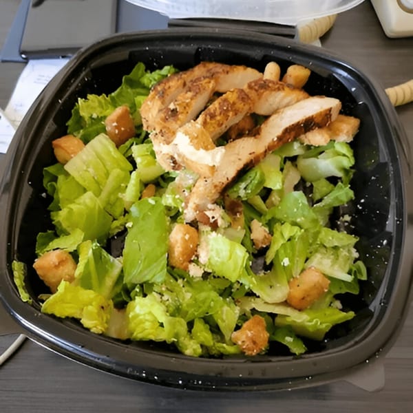 A Caesar salad with grilled chicken and croutons in a black to-go container from Who's Got Soul Southern Grill.